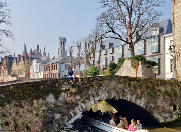Chris Heckmann and Nimarta Bawa sitting on a bridge in Bruges as a boat passes under